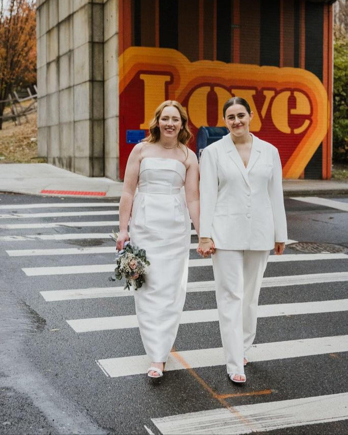 Two women in wedding attire standing on a crosswalk with a colorful 'LOVE' sign in the background.